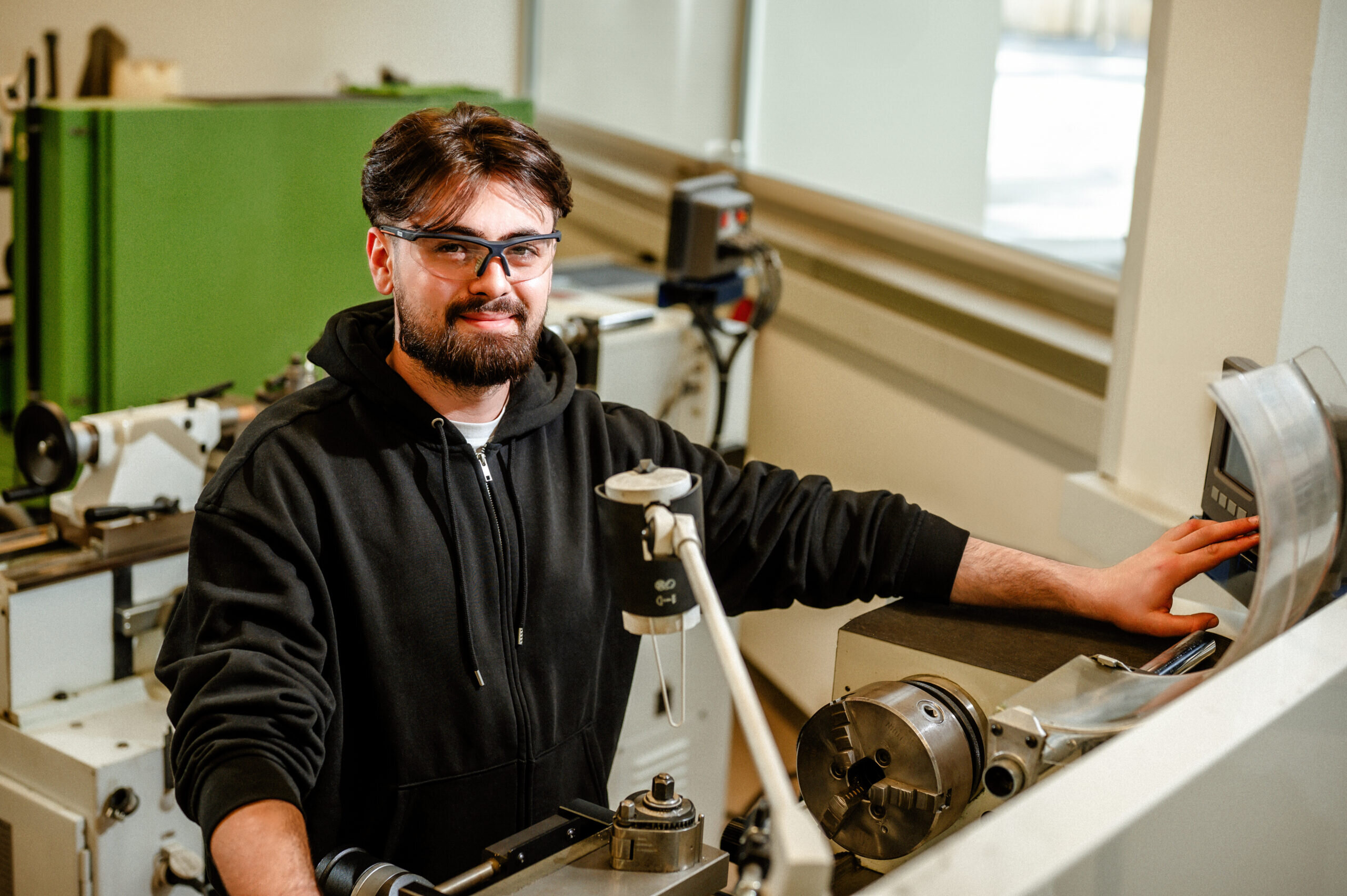 Ein junger Mann mit Brille lächelt am Arbeitsplatz in einer Maschinenwerkstatt. Er trägt einen schwarzen Hoodie und steht neben einer Drehmaschine.