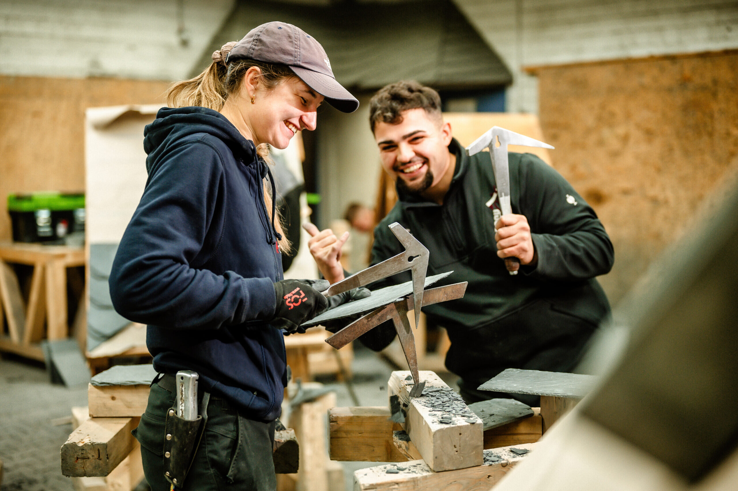 Zwei Personen arbeiten in einer Werkstatt mit Schiefer. Sie lächeln und zeigen sich gegenseitig ihre Werkzeuge. Teamarbeit und Freude am Handwerk!