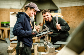 Zwei Personen arbeiten in einer Werkstatt mit Schiefer. Sie lächeln und zeigen sich gegenseitig ihre Werkzeuge. Teamarbeit und Freude am Handwerk!