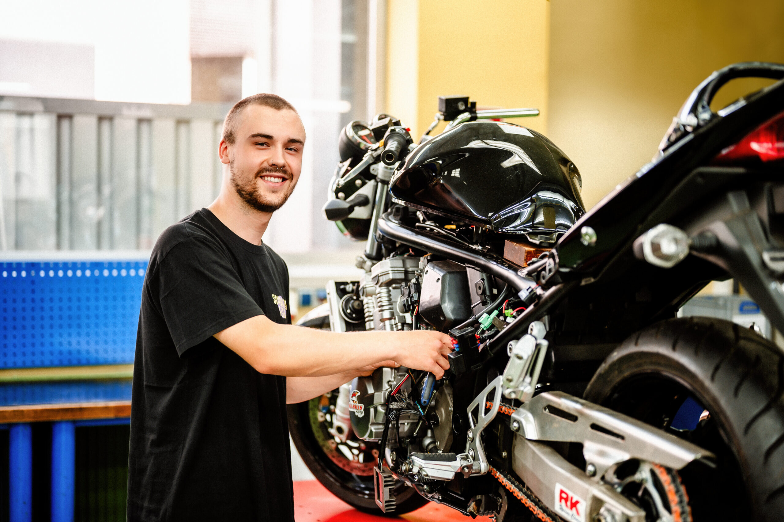 Ein junger Mann arbeitet an einem Motorrad und lächelt dabei. Im Hintergrund sind Werkzeuge und eine Werkbank zu sehen.