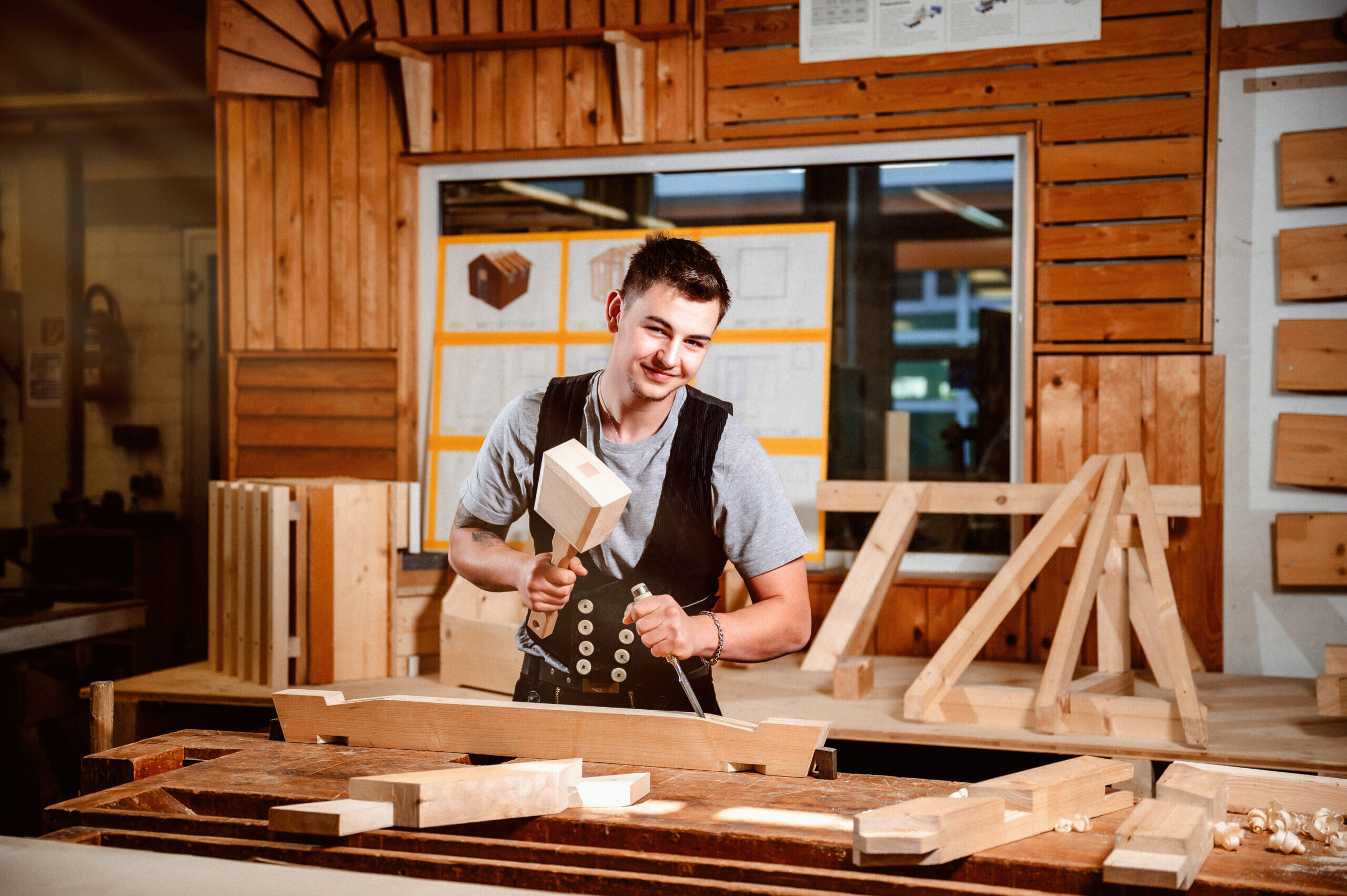 Der junge Mann arbeitet in einer Holzwerkstatt, konzentriert auf ein Projekt. Um ihn herum liegen verschiedene Holzstücke und Werkzeuge.