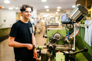 Ein junger Mann steht an einer Maschine in einer Werkstatt, lächelt und trägt eine Schutzbrille. Die Umgebung wirkt modern und gut beleuchtet.