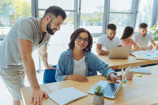 In einem modernen Büro arbeiten mehrere Menschen an Laptops. Eine Frau lächelt, während ein Mann ihr über die Schulter schaut.