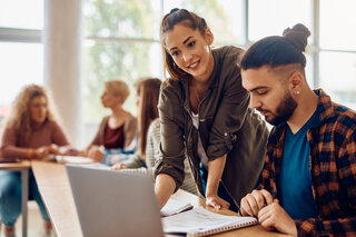 Zwei Personen arbeiten an einem Laptop in einem modernen Seminarraum. Eine Frau erklärt etwas, während der Mann aufmerksam zuschaut. Im Hintergrund sind weitere Personen zu sehen.