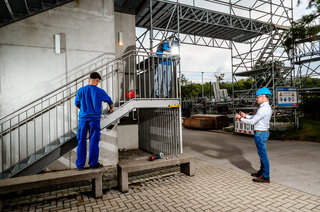 Zwei Arbeiter in Schutzkleidung begehen eine Baustelle. Einer arbeitet an der Treppe, während der andere Notizen macht. Sicherheitsmaßnahmen sind sichtbar.