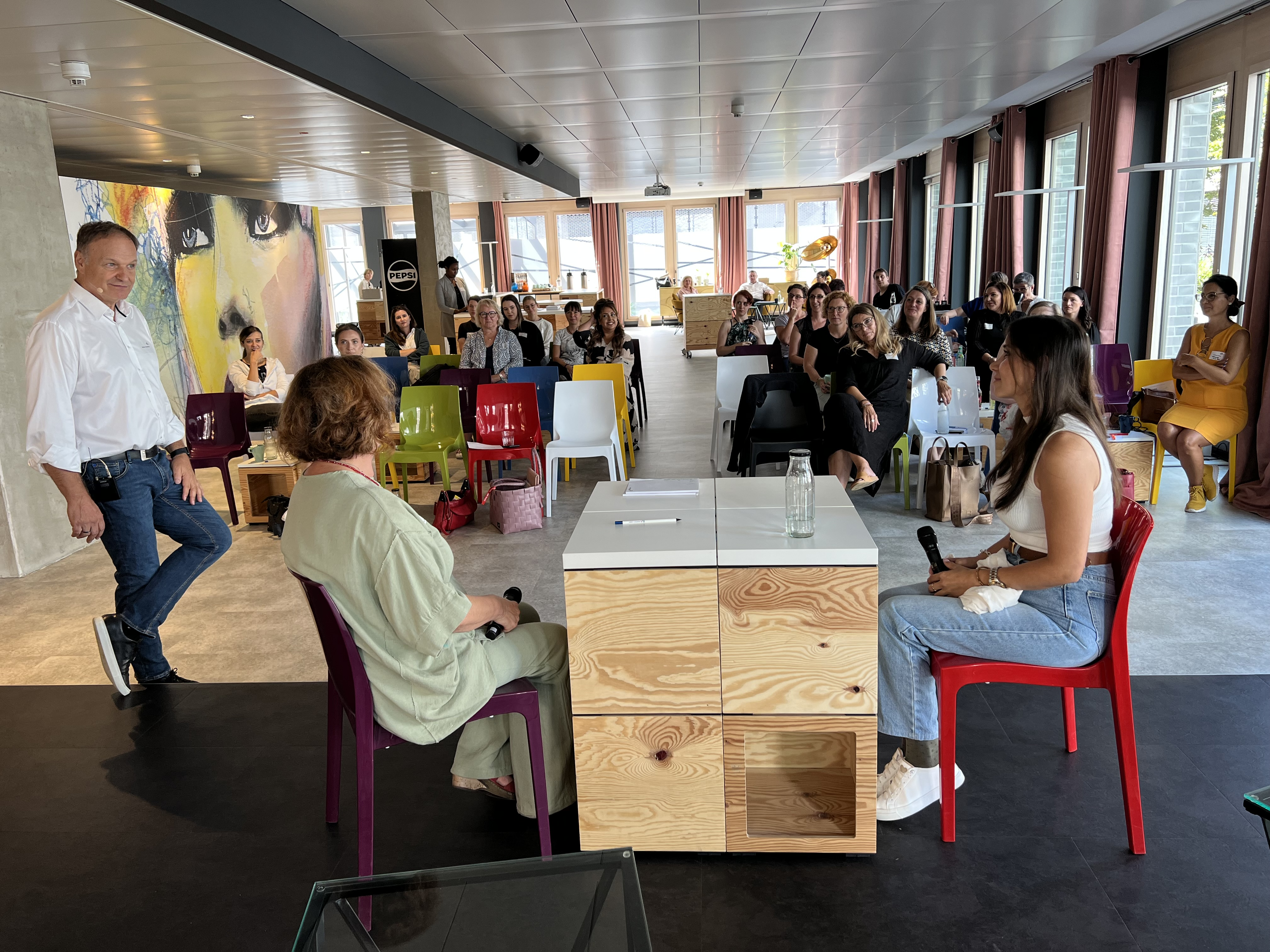 Das Bild zeigt eine Veranstaltung in einem modernen Raum. Zwei Frauen sitzen auf einem Podium, während ein Mann im Hintergrund spricht. Viele Zuhörer sind anwesend.