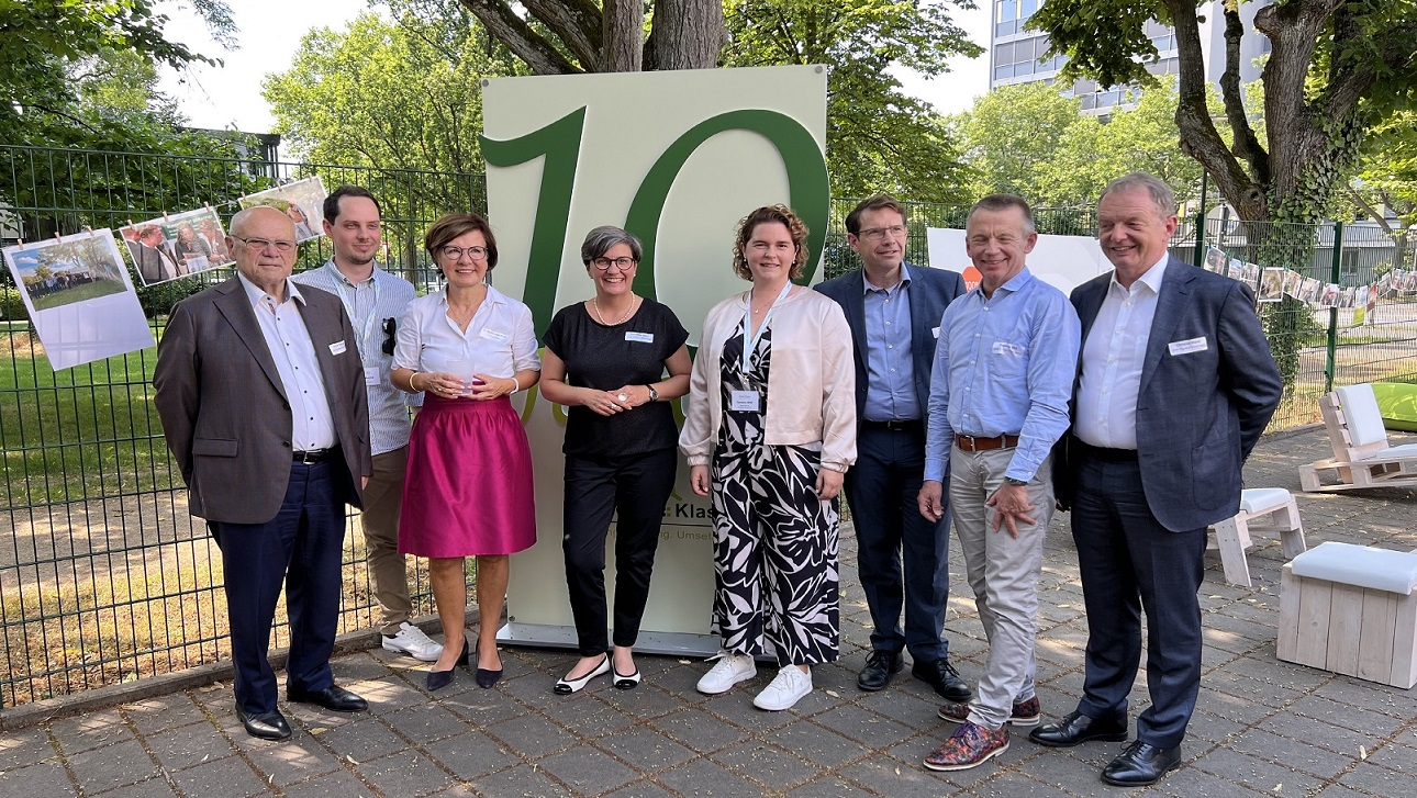 Auf dem Bild stehen mehrere Personen vor einer großen "10" in einer grünen Umgebung. Sie lächeln und scheinen an einem festlichen Anlass teilzunehmen.