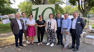 Auf dem Bild stehen mehrere Personen vor einer großen "10" in einer grünen Umgebung. Sie lächeln und scheinen an einem festlichen Anlass teilzunehmen.