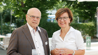 Das Bild zeigt ein älteres Paar, das draußen steht. Sie lächeln und halten transparente Becher in den Händen, umgeben von grüner Natur.